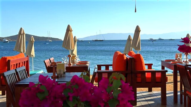 Vibrant outdoor restaurant with wooden furniture, bright orange cushions, and pink bougainvillea in the foreground, overlooking the Mediterranean Sea with yachts under a clear blue sky.