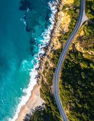 Aerial view of a coastal road winding along the edge. Turquoise waters crash against rocky shorelines and sandy beaches. Lush green vegetation flanks the path