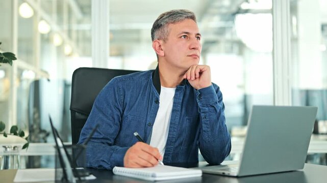 Thoughtful businessman in blue denim shirt over white t-shirt sits at modern office desk. Male brainstorms ideas, considering solutions, reflecting deep thought while working on laptop. Professional