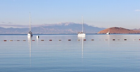 Two sailboats glide on calm Mediterranean waters, their masts reflected below a line of buoys, with a hazy mountain and arid island in the backdrop.