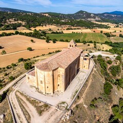 Aerial view of a large stone church atop a hill overlooking rural farmland and distant mountains under a sunny, cloud-filled sky
