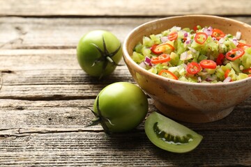 Tasty salsa in bowl and green tomatoes on wooden table, closeup. Space for text