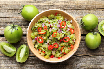 Tasty salsa in bowl and green tomatoes on wooden table, flat lay