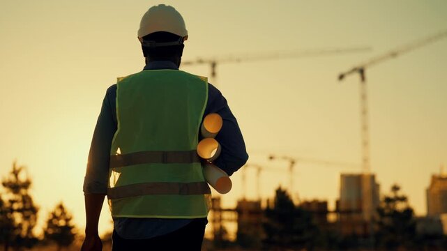 Safety precautions at construction site, back view of builder with white helmet. Professional foreman walking in sunset or sunrise time, city construction an development, cinematic shot, rear view