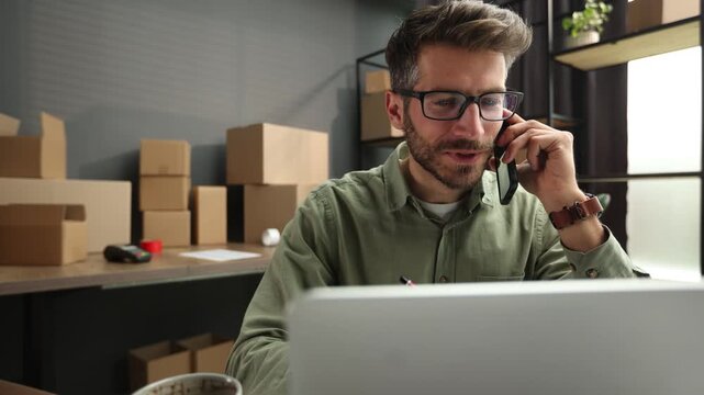 A man is talking on his cell phone while sitting at a desk with a laptop