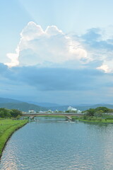 Wide river view with white arched bridge, manicured green lawns, distant mountains under dramatic blue sky with sunlit clouds at Dongshan Ecological Ark, Yilan.