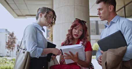Three diverse business partners in formal attire engage in a serious discussion outdoors. They are collaborating on important project details, exchanging ideas, and planning future strategies. - Powered by Adobe