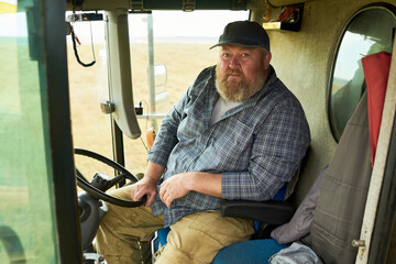 Portrait of middle aged Caucasian man sitting in tractor cab looking at camera, holding steering wheel with one hand, wearing cap and plaid shirt, rural landscape visible outside window