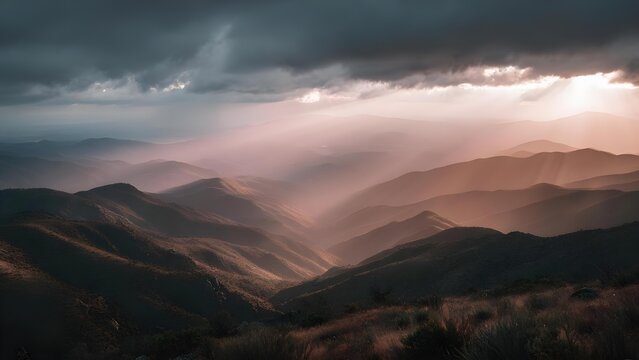 Sunbeams breaking through dark storm clouds illuminate pink-orange light over layered mountain ridges. Concept Dramatic mountain sunrise, Sunbeams through storm clouds, Pink-orange lighting