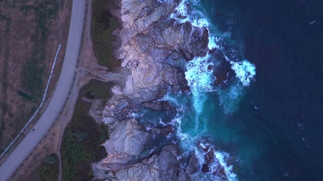 Aerial view of a circle of people sitting on the rocks of Beavertail State Park on Conanicut Island. The camera is high up, tilted down, boom down as the waves crash onto the rocky shore.