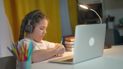 Child writes homework by laptop at desk. Headphones help kid during study. Girl learning education lessons. Focused student completes homework. Education setup for student with laptop and books - Powered by Adobe