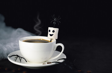 Hot coffee cup with smiling marshmallow ghost and spiders on dark background. Concept of festive morning coffee, Halloween treat, and fall mood. Copy space.