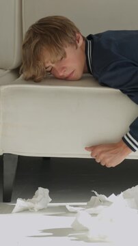 Teenage man lying on couch sick with flu, sneezing and reaching for tissues while surrounded by crumpled tissues, looking feverish, tired and miserable at home