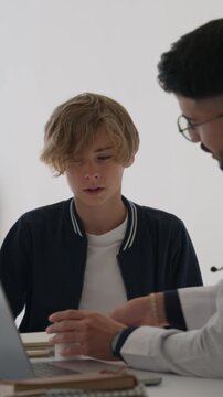 Youthful male physician in a white coat discusses diagnosis and treatment with a teenage patient at a modern clinic, using a laptop to explain medication and next steps