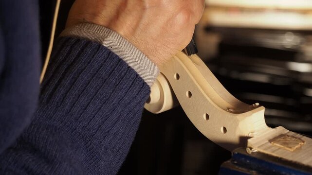 Craftsperson carving the scroll of a violin neck, using a small carving tool to refine the curves and details of the scroll head, the wood is securely held in a vise, allowing precise hand-tool work.