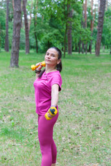 A happy mature woman in pink sportswear exercising with dumbbells in a green park, enjoying her workout.
