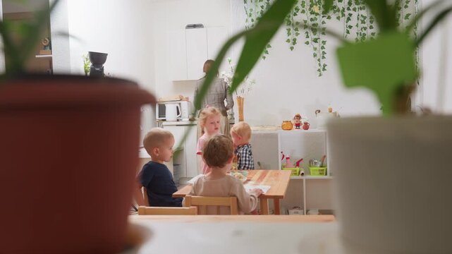 Scene in kindergarten viewed through decorative plants showing teacher near kitchen as girl in pink carries bowl with apple slices toward table while other children sit engaged