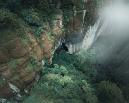 Dramatic aerial view of a lush green jungle canyon with a dark cave entrance and misty sunlight