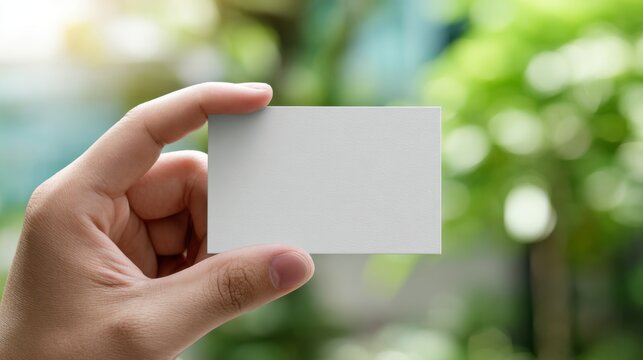 A close-up of a male hand holding a blank business card against a blurred green background. - Powered by Adobe