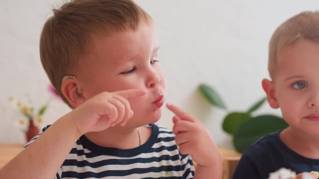 Close up of young boy with hearing aid gesturing with finger while expressing thought during meal beside another child in calm indoor setting background plants, and gentle focus depth