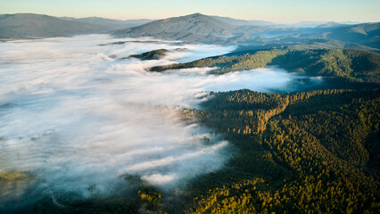 Stunning aerial view of sunrise over mountainous landscape, with thick fog blanketing valleys and hills. Sun's golden rays pierce through mist, casting serene glow over the entire scene.