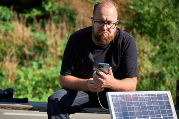 Fototapeta premium Bearded man using smartphone connected to photovoltaic solar panel. Integration of sustainable renewable energy into everyday life, demonstrating practical use of solar power for charging devices.