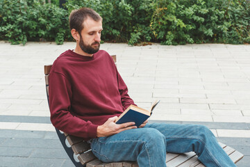 Obraz premium Young man with short hair and beard sitting on wooden bench outdoors, wearing a burgundy sweatshirt and blue jeans, reading book calmly, concept of relaxation, education, leisure and self-development