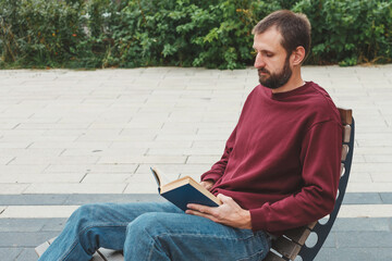 Obraz premium man with short hair and beard sitting on a wooden bench outdoors, wearing a burgundy sweatshirt and jeans, reading a book with concentration, concept of leisure, education, relaxation self-improvement