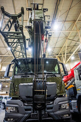 Front view of a powerful olive green heavy-duty truck equipped with a robust crane boom and lifting cables, showcased indoors. Represents strength, engineering, and industrial capability.