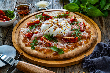 Pizza with cured ham, burrata cheese, tomatoes and arugula on wooden table