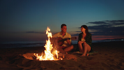 Young couple share an intimate moment by a glowing bonfire on a sandy beach at sunset, singing and playing guitar, enjoying a warm, peaceful evening together