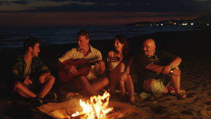 Four diverse friends are sitting on the sand around a crackling bonfire, a man playing guitar as they laugh and enjoy a fun night together by the ocean
