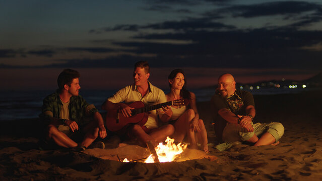 Diverse friends gathered around a glowing beach bonfire at dusk, singing, playing guitar and sharing laughter and stories while relaxing together by the ocean during summer vacation