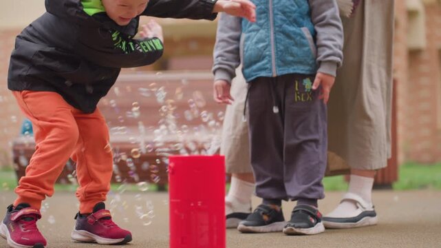 Excited boy in orange trousers bends hitting soap bubbles floating from red bubble machine while another child stands beside watching closely, playful outdoor scene filled with curiosity