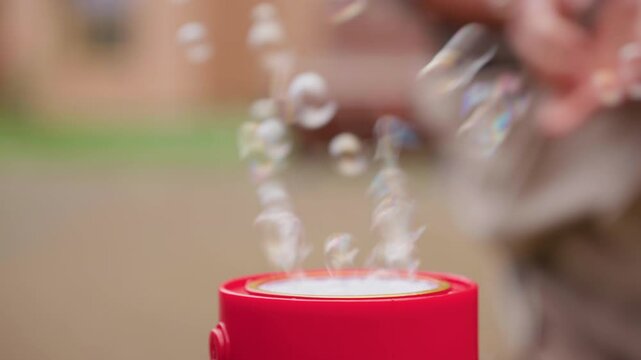 Close up of red bubble machine releasing clusters of delicate floating soap bubbles, blurred figure playing in background, soft focus captures movement