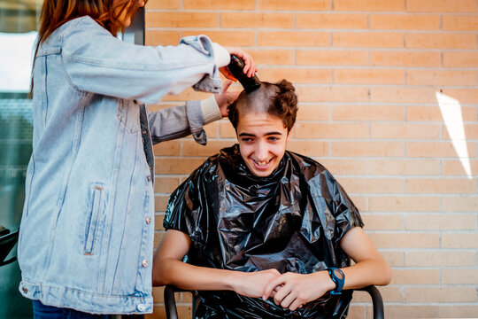 Mother shaving teenage son's head with electric razor against brick wall at home