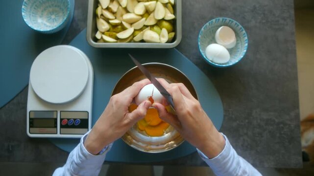 Woman's hands carefully breaking an egg with a knife into a metal bowl, preparing the batter for a delicious homemade pear charlotte with sliced pears and a kitchen scale on the table