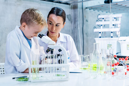 Mother scientist and son in lab coat using a microscope with guidance from a female scientist in a modern laboratory, surrounded by glassware and equipment, highlighting STEM education and research.