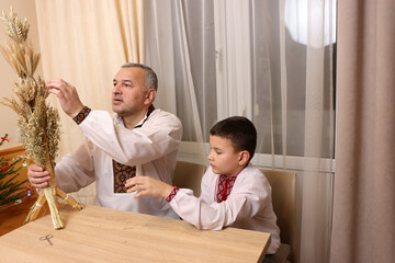 Fototapeta premium Father and son in embroidered shirts examine didukh made of wheat stalks, symbol of prosperity.
