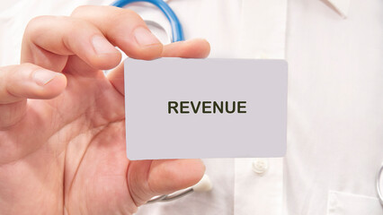 A person holds a card with the word revenue prominently displayed, set against a backdrop of healthcare tools, capturing the essence of financial discussions in medical environments