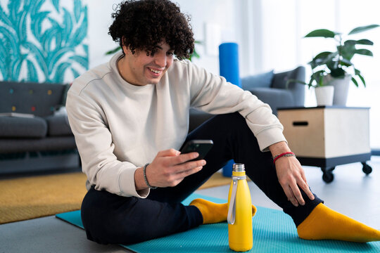 Smiling young Arab male in activewear watching online tutorial on smartphone while drinking water from bottle during fitness training at home