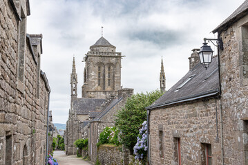 A picturesque stone house sits beside a lush garden filled with bright hydrangeas along a cobblestone street in the quaint historic village of Locronan in Brittany, France.