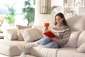 Happy woman in winter at home reading a book drinking coffee