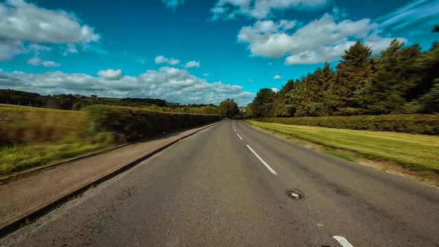 Driving along countryside road surrounded by green fields and trees under bright blue sky POV. Moving through rural asphalt road bordered by hedges and forest first person view. Traveling down scenic