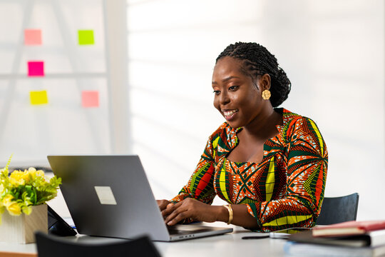 African woman wearing vibrant ankara print dress uses laptop for remote work, online learning, video calls, online shopping, digital banking, social media and internet browsing - Powered by Adobe