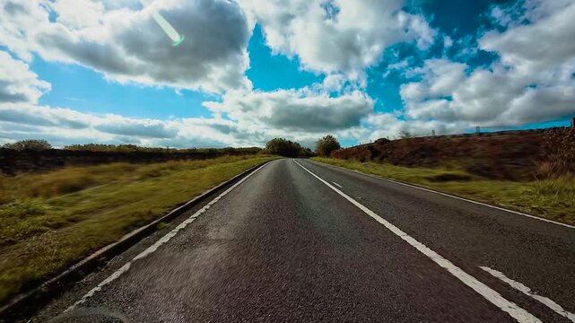 Car driving through countryside road with left hand traffic in England surrounded by open landscape POV. SUV moving along rural British highway bordered by grass and bushes under bright sky first