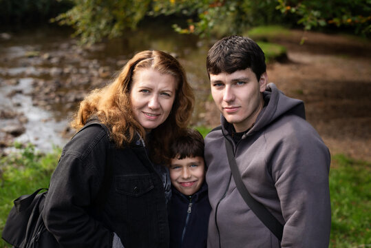 A warm outdoor family portrait featuring a mother, eldest son, and young son close together by a riverbank. They are chatting, smiling and happy to be together again after long separation