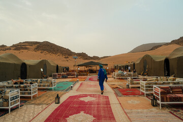 Cluster of traditional tents arranged in the Sahara desert, Moroccan carpets and sofas laid out, a local man stands centrally amid the rugged red-toned landscape of Morocco.