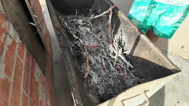 High-angle, textured detail of dry wood and small branches nestled into a bed of white ash in a metal container, ready for lighting the perfect charcoal fire.