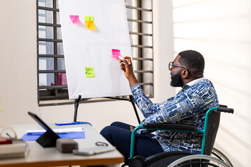 African male entrepreneur in a wheelchair strategizing with sticky notes on a whiteboard, inclusion...